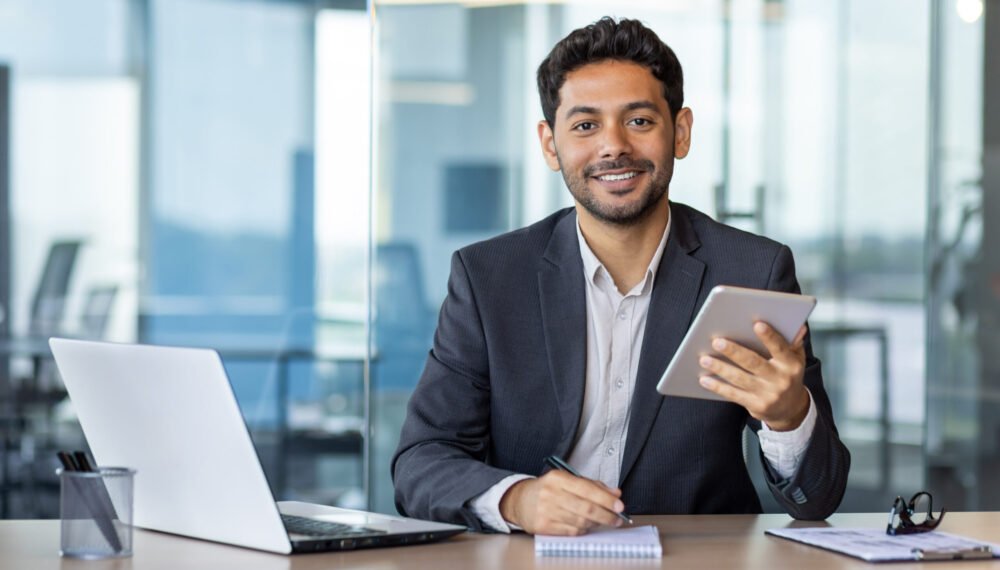 Portrait of young businessman with tablet computer inside office, hispanic man smiling and looking at camera, working with laptop, using app on tablet.