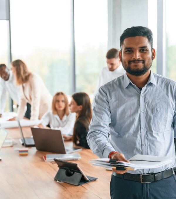Indian man is standing in front of colleagues that are using laptop. Group of office workers are together indoors.